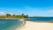 Une étroite bande de sable doré formant une plage incurvée, reliant le continent à une petite île verdoyante à Saint-Briac-sur-Mer, sur la Côte d'Émeraude près de Saint-Malo, avec l'océan turquoise s'étendant à l'horizon sous un ciel bleu.