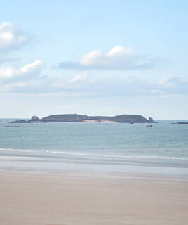 Une petite île rocheuse au loin, l'Île de Cézembre près de Saint-Malo, émergeant des eaux calmes de l'océan, vue depuis une plage de sable sous un ciel partiellement nuageux.