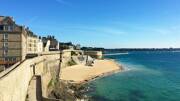 Les Remparts de Saint-Malo, en Bretagne, longeant une plage de sable fin et l'eau turquoise de la mer, avec les maisons de la vieille ville et les tours fortifiées sous un ciel bleu.