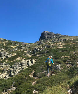 Une randonneuse avec un sac à dos et des bâtons de marche gravit un sentier rocailleux et verdoyant dans le Massif de la Clape, près de Saint-Pierre-la-Mer, sous un ciel bleu clair.