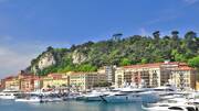 Vue sur le port, avec ses yachts de luxe amarrés et ses bâtiments colorés le long du quai, adossés à une colline verdoyante sous un ciel bleu intense.