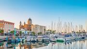 Le port de Saint-Raphaël avec ses nombreux bateaux de plaisance et voiliers amarrés, reflétés dans l'eau calme, sous un ciel clair et la silhouette de la Basilique Notre-Dame-de-la-Victoire en arrière-plan.