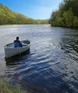 Un homme seul dans une petite barque sur le lac de Sanguinet, au milieu des vagues, avec des rives boisées en arrière-plan sous un ciel bleu clair.