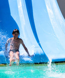 Un enfant souriant et éclaboussé sort d'un toboggan aquatique bleu, atterrissant dans une piscine, probablement dans un camping près de Sanguinet.