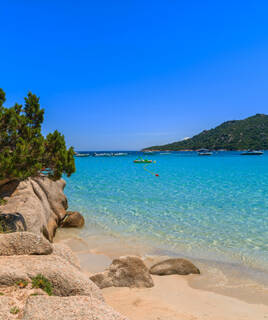 La plage de Santa Giulia en Corse, avec son sable blanc, ses eaux turquoise et cristallines, bordée de rochers et de végétation méditerranéenne, avec des bateaux au large et des collines en arrière-plan sous un ciel bleu éclatant.
