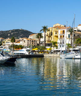 Le port de Porto-Vecchio, près de Santa Giulia en Corse, avec ses bateaux amarrés et ses bâtiments colorés le long du quai, bordé de palmiers sous un ciel bleu clair.