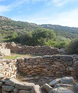 Les ruines de l'ancien site de Ceccia, près de Santa Giulia en Corse, avec des murs de pierre sèche et des vestiges archéologiques, au milieu d'une végétation méditerranéenne luxuriante et de collines sous un ciel bleu clair.