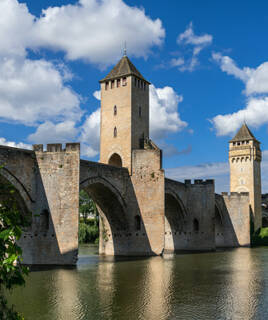 Le Pont Valentré à Cahors, près de Sarlat, un pont médiéval fortifié avec ses trois tours distinctives enjambant la rivière Lot, sous un ciel bleu parsemé de nuages blancs.