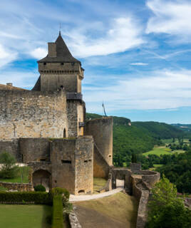 Le Château de Castelnaud, près de Sarlat en Dordogne, une forteresse médiévale en pierre avec ses tours et remparts, surplombant une vallée verdoyante sous un ciel bleu parsemé de nuages blancs.