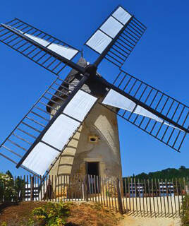 Un moulin à vent traditionnel avec des voiles blanches, visible au Parc du Bournat près de Sarlat, sous un ciel bleu clair, entouré d'une petite clôture en bois.