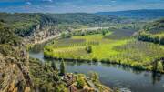 Le village de La Roque-Gageac, près de Sarlat en Dordogne, s'étire le long de la rivière Dordogne au pied d'une falaise, avec des champs et des forêts verdoyantes en arrière-plan sous un ciel bleu parsemé de nuages.