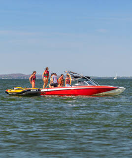 Un groupe de personnes en gilets de sauvetage se préparant pour des activités nautiques sur un bateau à moteur rouge, avec une bouée jaune à proximité, sur les eaux du Golfe du Morbihan à Sarzeau, sous un ciel bleu clair.