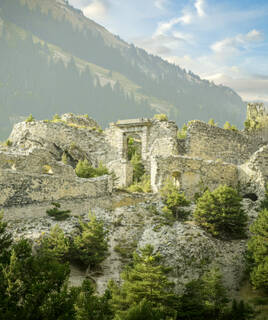 Les ruines d'une forteresse médiévale, probablement le fort d'Esseillon à Aussois en Savoie, perchées sur une colline rocheuse et boisée avec des montagnes en arrière-plan sous un ciel partiellement nuageux.