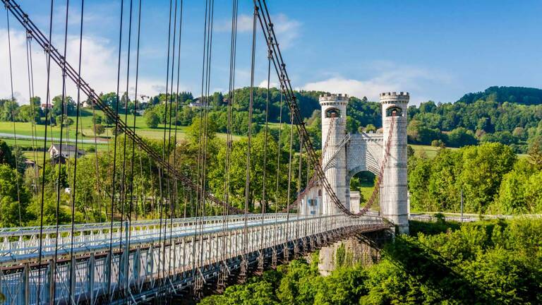 Le Pont de la Caille, un pont suspendu emblématique près de Chambéry en Savoie, avec ses tours en pierre et ses câbles, enjambant une vallée verdoyante sous un ciel bleu.
