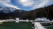 Le Lac de la Rosière à Courchevel, en Savoie, entouré de forêts de pins enneigées et de montagnes imposantes sous un ciel bleu avec des nuages, avec un ponton enneigé sur l'eau.