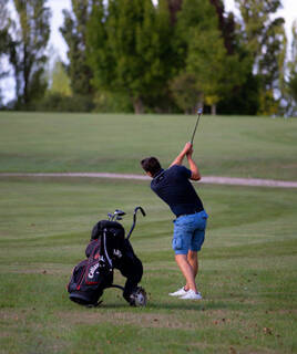 Un golfeur en plein swing sur le parcours de golf de Seignosse, avec son sac de clubs posé à côté de lui sur le gazon vert, sous un ciel partiellement couvert.