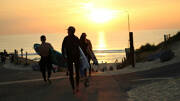 Des surfeurs portant leurs planches marchent sur un chemin menant à la plage d'Hossegor, près de Seignosse, au lever ou au coucher du soleil, sous un ciel orangé.