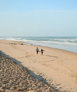 Deux surfeurs marchant sur la vaste plage de sable de Seignosse, portant leurs planches vers l'océan Atlantique agité, sous un ciel bleu clair.