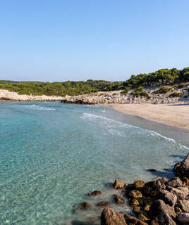 Une crique isolée avec une plage de sable fin et des eaux cristallines turquoise, bordée de rochers et de pins méditerranéens, typique du littoral varois près de La Seyne-sur-Mer, sous un ciel bleu clair.