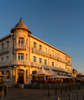 Un élégant bâtiment ancien avec une tourelle, abritant des commerces et une terrasse de restaurant animée dans le centre historique de Soulac-sur-Mer, éclairé par la lumière chaude du soleil couchant.