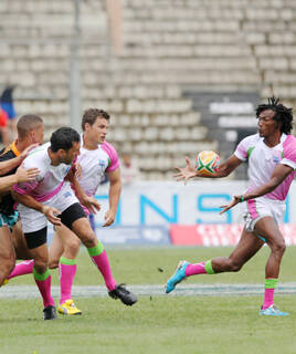 Un match de rugby à Toulon, avec des joueurs en action sur le terrain, certains portant des maillots roses et blancs, sous un ciel partiellement couvert et des tribunes en arrière-plan.