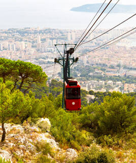 Une cabine rouge du téléphérique du Mont Faron à Toulon, descendant au-dessus d'une végétation dense, offrant une vue panoramique sur la ville et la mer en arrière-plan sous un ciel clair.