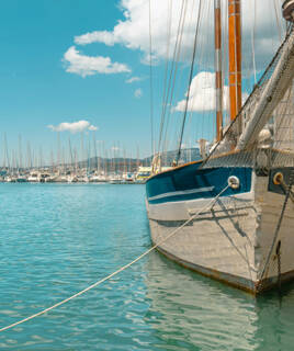 Un voilier classique blanc et bleu amarré dans le port de Toulon, avec de nombreux autres bateaux en arrière-plan et des collines, sous un ciel bleu parsemé de nuages blancs.