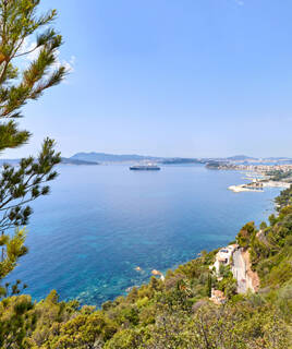 Une vue panoramique depuis le Sentier du Littoral à Toulon, surplombant la Méditerranée avec un grand paquebot au large, le port et la ville en arrière-plan, encadrée par des arbres et de la végétation luxuriante sous un ciel bleu clair.