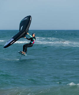 Une personne pratiquant le wingfoil sur les eaux de Valras-Plage, glissant sur une planche avec une aile gonflable, sous un ciel clair, avec la ligne d'horizon lointaine en arrière-plan.