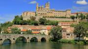 La ville de Béziers, près de Valras-Plage, avec sa cathédrale Saint-Nazaire perchée sur une colline dominant la rivière Orb et son pont en pierre, sous un ciel bleu parsemé de nuages blancs.