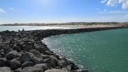 Le Cap d'Agde, près de Valras-Plage, avec une digue rocheuse s'avançant dans la mer aux eaux turquoise, et une plage de sable animée avec des bâtiments en arrière-plan sous un ciel bleu parsemé de nuages blancs.