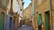 Une rue pavée étroite du centre historique de Pézenas, près de Valras-Plage, bordée de bâtiments anciens en pierre avec des volets colorés sous un ciel bleu lumineux.