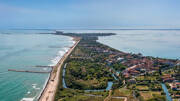 Vue aérienne du Lido de Venise, une étroite bande de terre entre l'Adriatique et la lagune de Venise, avec des plages, des jetées, des zones vertes et des bâtiments, sous un ciel clair.