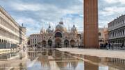 La Place Saint-Marc à Venise, inondée d'eau reflétant la Basilique Saint-Marc et le Campanile, avec des touristes dispersés et des bâtiments historiques sous un ciel partiellement nuageux.