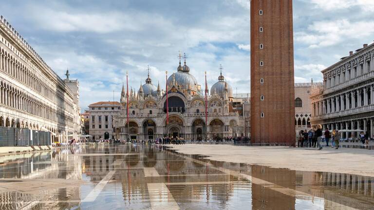 La Place Saint-Marc à Venise, inondée d'eau reflétant la Basilique Saint-Marc et le Campanile, avec des touristes dispersés et des bâtiments historiques sous un ciel partiellement nuageux.