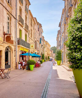 Une rue piétonne animée du centre historique de Vias, bordée de bâtiments anciens aux façades en pierre, de terrasses de cafés colorées avec des parasols jaunes et de grands pots de fleurs verts, sous un ciel clair.