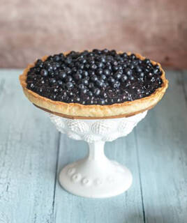 Une tarte généreusement garnie de myrtilles fraîches, posée sur un présentoir blanc en dentelle, évoquant les spécialités vosgiennes à déguster.