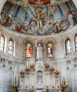 Intérieur de la Basilique du Bois-Chenu à Domrémy-la-Pucelle, révélant son autel orné de bougies et une statue de Jeanne d'Arc sous une coupole aux fresques détaillées et des vitraux colorés.