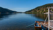 Vue panoramique du Lac de Gérardmer avec une barque en bois amarrée à un ponton au premier plan, entouré de montagnes verdoyantes sous un ciel clair.