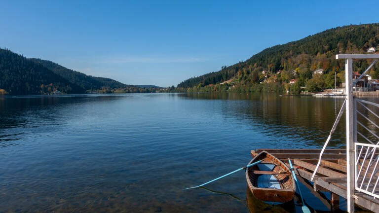 Vue panoramique du Lac de Gérardmer avec une barque en bois amarrée à un ponton au premier plan, entouré de montagnes verdoyantes sous un ciel clair.