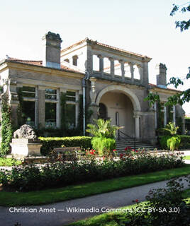 Une élégante maison de style romain avec une façade en briques claires et des colonnes, entourée de jardins fleuris et de statues, visible au Musée d'Art Contemporain de la ville d'Épinal, dans les Vosges, sous un ciel clair.