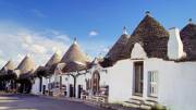 Une rangée de trulli, maisons traditionnelles aux toits coniques en pierre, dans le village d'Alberobello dans les Pouilles, avec des boutiques le long de la rue sous un ciel bleu parsemé de nuages.