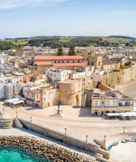 Une vue aérienne du Château d'Otrante dans les Pouilles, une imposante forteresse en bord de mer, entourée de la ville historique aux toits de tuiles, avec un port et des eaux turquoise, sous un ciel bleu clair.