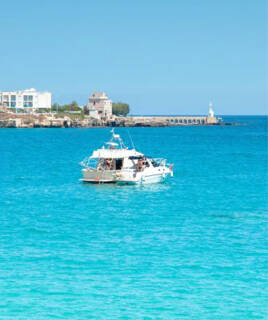 Un bateau de plaisance blanc flottant sur les eaux turquoise de la côte des Pouilles, avec un port et un phare en arrière-plan sous un ciel bleu éclatant, invitant à une excursion en bateau.
