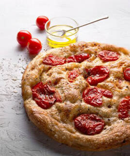 Une focaccia Barese, spécialité des Pouilles, avec des tomates cerises et des herbes sur sa surface dorée, accompagnée de tomates fraîches et d'huile d'olive en arrière-plan.