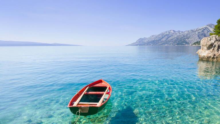Une petite barque rouge flottant sur les eaux cristallines de la mer Adriatique dans les Pouilles, avec des montagnes et un ciel bleu clair en arrière-plan.