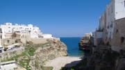 Polignano a Mare dans les Pouilles, avec ses maisons blanches perchées sur des falaises rocheuses surplombant une crique aux eaux turquoise, et la mer Adriatique s'étendant à l'horizon sous un ciel bleu clair.