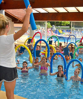 Un groupe d'enfants et un adulte participant à une activité aquatique dans une piscine couverte, utilisant des frites de piscine et des planches de natation, avec un toboggan visible en arrière-plan.