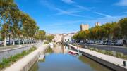 Canal de la ville de Narbonne, avec un petit bateau, une allée d'arbre de chaque côté du canal, avec une vue de la ville en fond sous un ciel bleu