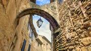 Maison en pierre de couleur claire avec une arche ancienne, une lanterne noire accroché à l'arche, sous un ciel bleu